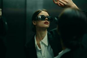 stilvoll Frau im ein schwarz Jacke und Sonnenbrille posieren im ein Aufzug Aufzug in der Nähe von das Spiegel, Mode Modell, dunkel filmisch Licht und Farbe Matrix, Glanz Jahrgang foto