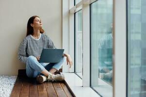 Geschäft Frau Sitzung mit ein Laptop beim das Fenster in voller Länge und lächelnd und suchen aus das Fenster, glücklich Frau Arbeit online im das Büro, Kopieren Raum foto