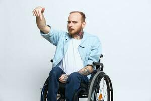 ein Mann im ein Rollstuhl und zeigen ein Finger bei, Kopieren Raum, mit Tätowierungen auf seine Hände sitzt auf ein grau Studio Hintergrund, Gesundheit Konzept ein Person mit Behinderungen, ein echt Person foto