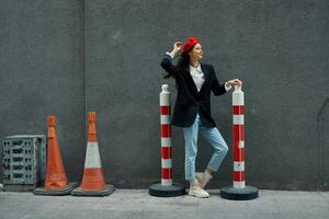 Mode Frau Stehen gelehnt gegen ein Mauer Straße gegen ein Hintergrund von das Stadt Straße funktioniert Tourist im stilvoll Kleider mit rot Lippen und rot Baskenmütze, reisen, filmisch Farbe, retro Jahrgang Stil. foto