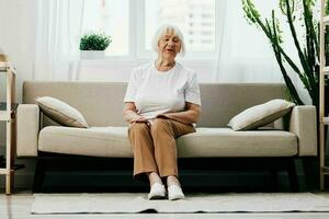 Alten Frau sitzt auf Sofa beim heim, hell geräumig Innere im alt Alter lächeln, Lebensstil. Oma mit grau Haar im ein Weiß T-Shirt und Beige Hose. foto