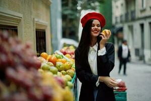 Frau Lächeln mit Zähne Tourist Spaziergänge im das Stadt Markt mit Früchte und Gemüse wählen Waren, stilvoll modisch Kleider und bilden, Frühling gehen, Reise. foto