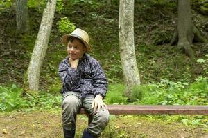 komisch kaukasisch Junge im ein Reisende Hut und Windjacke sitzt auf ein Bank foto