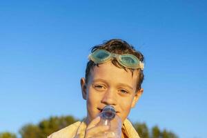 glücklich Junge im Schwimmen Brille Getränke Wasser von ein Flasche auf das Strand foto