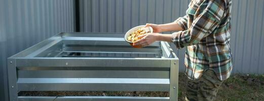 ein Frau im ein Plaid Hemd gießt Essen Abfall von ein Schüssel in ein Kompost Haufen von Kartoffel und Karotte Schalen. Kompost Box gemacht von Metall, umweltfreundlich Dünger zum das Garten foto