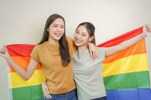 glücklich lesbisch, schön asiatisch jung zwei Frauen, Mädchen Fröhlich, Paar Liebe Moment Ausgaben gut Zeit zusammen, halten oder winken lgbt Regenbogen, Stolz Flagge auf isoliert Weiß Mauer Hintergrund zusammen beim heim. foto