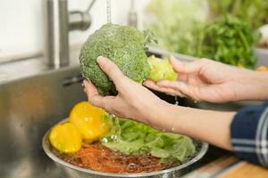 schließen oben asiatisch jung Frau Waschen Brokkoli, Tomate, Karotte frisch Gemüse, Paprika mit Spritzen Wasser im Becken von Wasser auf sinken im Küche, vorbereiten frisch Salat, Kochen Mahlzeit. gesund Essen Personen. foto