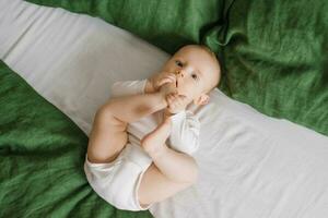 süß Baby ist Lügen auf ein Grün und Weiß Tagesdecke auf das Bett. ein lächelnd Kind haben seine Füße im ein sonnig Schlafzimmer. foto