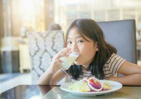 asiatisch wenig süß Mädchen ist Trinken ein Glas von Milch und Essen Salat foto