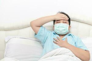 asiatisch Fett Junge tragen chirurgisch Maske mit ein hoch Fieber und Truhe Schmerzen auf Bett, foto