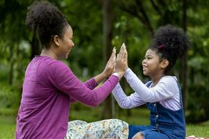 Die afroamerikanische Mutter lacht, während sie mit ihrer kleinen Tochter Patty Cake spielt, während sie ein Sommerpicknick im öffentlichen Park für das Konzept des Wohlbefindens und des Glücks hat foto