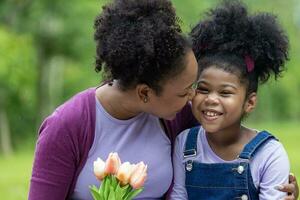 afrikanisch Mutter halten Strauß von Tulpe Blumen und das Tochter küssen jeder andere während glücklich genießen Picknick im das Öffentlichkeit Park während Sommer- zum Familie Liebe und Pflege im Mutter Tag Feier foto