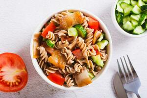 schließen oben von herzhaft Salat von Pasta, Kirsche Tomaten, Gurken und gebraten Speck im ein Schüssel oben Aussicht foto