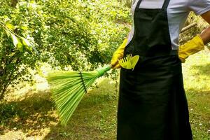 ein Frau im ein Schürze und Haushalt Handschuhe mit Garten Werkzeuge im ihr Tasche hält ein Besen im ihr Hände, im das Garten im Sommer- foto