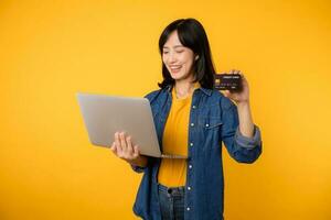 glücklich jung Frau von asiatisch ethnische Zugehörigkeit tragen Gelb T-Shirt Denim Hemd mit Laptop pc Computer halt Anerkennung Bank Karte Einkaufen online bestellen Lieferung isoliert auf einfach Gelb Hintergrund. foto