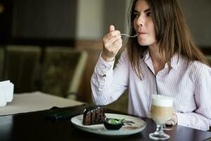 jung schön Brünette Frau sitzen im Kaffee Geschäft Cafe Restaurant drinnen und Essen Schokolade Brownie Dessert Kuchen, Gabel in der Nähe von das Mund. foto