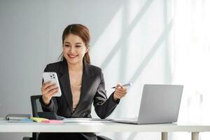 attraktiv Brünette asiatisch Frau mit Handy, Mobiltelefon Telefon, Lächeln und glücklich während Sitzung beim Arbeiten Schreibtisch im modern Büro. foto