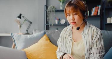 Asiatische Geschäftsfrau, die Laptop verwendet, spricht mit Kollegen über den Plan im Videoanruf, während sie vom Haus im Wohnzimmer arbeitet