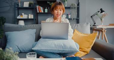 Asiatische Geschäftsfrau, die Laptop verwendet, spricht mit Kollegen über den Plan im Videoanruf, während sie vom Haus im Wohnzimmer arbeitet