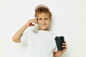 Foto Porträt lockig wenig Junge im ein Weiß T-Shirt Deckel mit ein Telefon im ein Glas mit ein trinken Licht Hintergrund unverändert