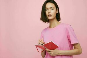 jung Frau im ein Rosa T-Shirt rot Notizbuch und Stift Rosa Hintergrund foto