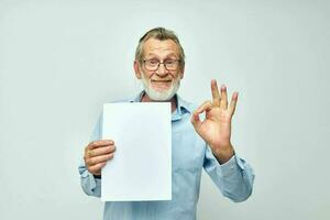 Senior grauhaarig Mann im ein Blau Hemd und Brille ein Weiß Blatt von Papier abgeschnitten Aussicht foto