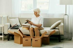 Alten Frau sitzt auf ein Sofa beim Zuhause mit Boxen. Sammeln Dinge mit Erinnerungen Alben mit Fotos und Foto Frames ziehen um zu ein Neu Platz Reinigung Dinge und ein glücklich lächeln. Lebensstil Ruhestand.