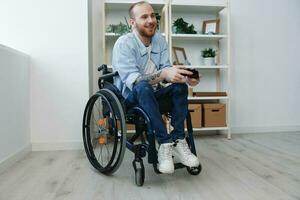 ein Mann im ein Rollstuhl Spieler Theaterstücke Spiele mit ein Joystick im seine Hände beim heim, Kopieren Raum, mit Tätowierungen auf seine Hände, Gesundheit Konzept Mann mit Behinderungen, echt Person foto