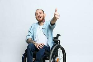 ein Mann im ein Rollstuhl sieht aus beim das Kamera zeigt an ein Daumen hoch, Glück, mit Tätowierungen auf seine Hände sitzt auf ein grau Studio Hintergrund, Gesundheit Konzept Mann mit Behinderungen foto