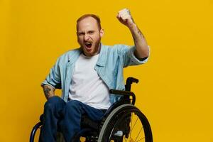 Mann im ein Rollstuhl Geschrei wütend, zeigt an Faust sieht aus beim das Kamera ist nicht erfreut, mit Tätowierungen auf seine Hände sitzt auf Gelb Hintergrund von das Studio, das Konzept von Gesundheit ist Person mit Behinderungen foto