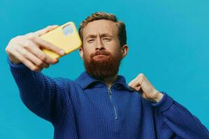 Porträt von ein Mann mit ein Telefon im seine Hände Blogger nimmt Selfies, auf ein Blau Hintergrund. kommunizieren online Sozial Medien, Lebensstil foto