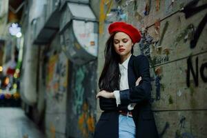 Mode Frau Porträt Gehen Tourist im stilvoll Kleider mit rot Lippen Gehen Nieder ein eng Stadt Straße, reisen, filmisch Farbe, retro Jahrgang Stil, dramatisch gegen ein Mauer mit Graffiti. foto