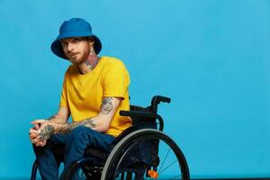 ein Mann im ein Rollstuhl Probleme mit das Bewegungsapparat System sieht aus beim das Kamera im ein T-Shirt mit Tätowierungen auf seine Waffen sitzt auf ein Blau Studio Hintergrund, voll Leben, echt Person, Gesundheit Konzept foto
