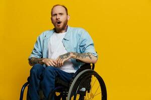 ein Mann im ein Rollstuhl überrascht, mit Tätowierungen auf seine Waffen sitzt auf ein Gelb Studio Hintergrund, das Konzept von Gesundheit ist ein Person mit Behinderungen foto