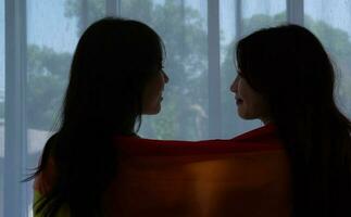 das lgbt Paar saß auf das Bett, bedeckt im Regenbogen Flaggen, spähen aus das Fenster zu beobachten das Natur im das Hotel Zimmer. foto