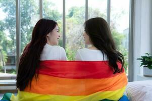 das lgbt Paar saß auf das Bett, bedeckt im Regenbogen Flaggen, spähen aus das Fenster zu beobachten das Natur im das Hotel Zimmer. foto