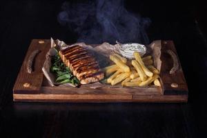 Lachsfiletsteak mit Pommes Frites auf einem Holztablett, schöne Portion, dunkler Hintergrund foto