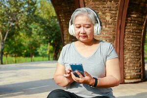 asiatisch Alten Frau ausüben im das Park im das Morgen sie sitzt und entspannt, halten ihr Smartphone und tragen Kopfhörer zu Hör mal zu zu Musik. Alten Gesundheit Pflege Konzept foto
