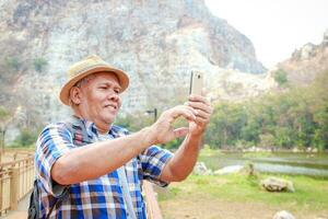 asiatisch Alten Mann Reisen im Natur Stehen mit ein Smartphone zu nehmen Bilder von das felsig Berg Landschaft. Reise Konzept, glücklich Pensionierung Leben, Gesundheit Pflege foto