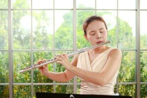 asiatisch Frau tragen ein Sahne farbig Kleid sie ist spielen das Flöte, ein International Wind Instrument. abspielen klassisch Lieder. durchführen Konzerte spielen klassisch Musik- im ein Band. Musik- Lernen foto