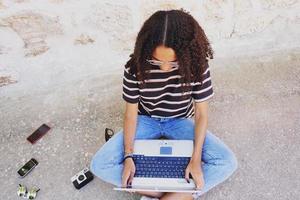 ein Porträt einer fokussierten jungen schwarzen Frau mit lockigem Haar, die eine Brille, Jeans und ein gestreiftes T-Shirt trägt, neben Technologie wie Smartphones und Kameras, die auf dem Boden sitzen und arbeiten oder Hausaufgaben machen foto