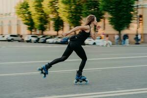 professionelle weibliche roller demonstriert ihre fähigkeiten von rollerblading fahrten sehr schnell auf der straße entlang der stadt genießt sonnigen tag in schwarzer aktiver kleidung. aktives Lifestyle-Hobby und Fitness-Aktivität foto