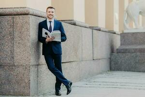 erfolgreich Boss bereitet vor zum Präsentation, hält Zeitschrift, liest, steht durch Büro Gebäude, formal Kleidung foto