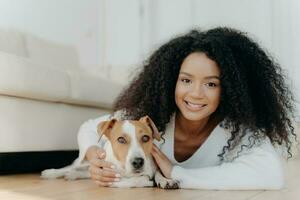 ziemlich Mädchen mit afro Haar Lügen auf Leben Zimmer Boden, Teilen angenehm Emotionen mit neu angenommen Hund. Geliebte Haustier im Neu Wohnung, Erstellen schön Momente zusammen. foto