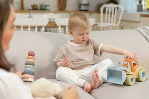 glücklich Familie beim heim. Mutter und Baby Junge spielen mit Spielzeuge im Couch beim Zuhause drinnen. wenig Kleinkind Kind und Babysitter Tagesmutter haben Spaß zusammen. jung Frau Mama Kind Sohn sich ausruhen im Leben Zimmer. foto