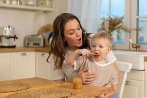 glücklich Familie beim heim. Mutter Fütterung Baby im Küche. wenig Junge mit unordentlich komisch Gesicht isst gesund Lebensmittel. Kind lernt Essen durch selbst halten Löffel. Frau Mama geben Essen zu Kind Sohn. selbst füttern. foto