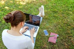 junge Frau, die auf grünem Rasen im Stadtpark sitzt und an Laptop-PC-Computer arbeitet. freiberufliches Geschäftskonzept foto