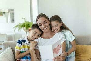 froh jung Mutter und wenig Kinder Sitzung auf Sofa und Umarmen während lesen wünscht sich und Herzliche Glückwünsche mit Urlaub im vorgestellt Postkarte beim Zuhause foto