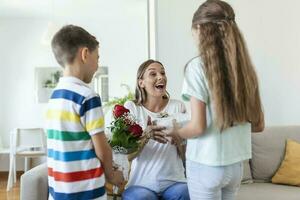 glücklich Kinder geben Geschenk ein Blumen zu Mutter. glücklich Mütter Tag. Kinder Junge und Mädchen gratulieren lächelnd Mutter, geben ihr Blumen Strauß von Rosen und ein Geschenk Box während Urlaub Feier foto