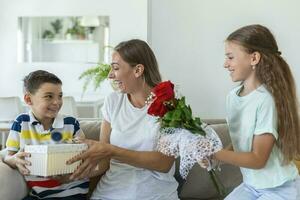 heiter wenig Mädchen mit Strauß von Rosen Blumen und jüngste Bruder mit Geschenk Box lächelnd und Glückwunsch glücklich Mama auf Mutter Tag beim heim. glücklich Mütter Tag foto